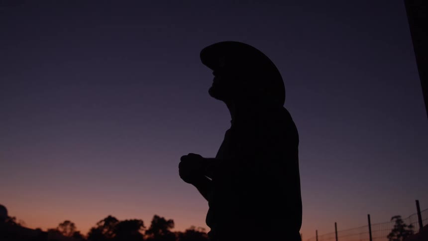The dark silhouette of a cowboy in a hat stands holding his hands together in prayer or reflection against a colorful twilight sky