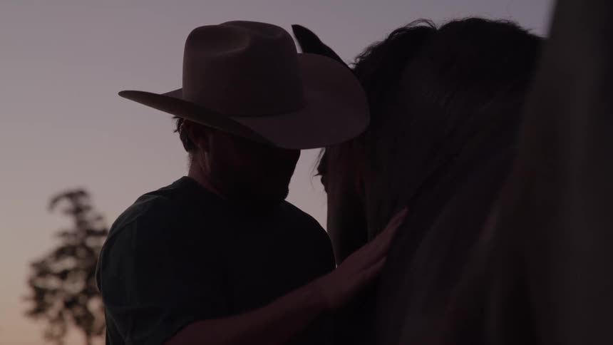 A middleaged man gently pets a young colt in a ranch at sundown bathed in the warm glow of fading light