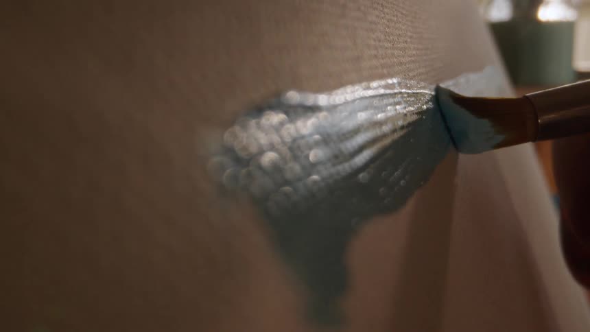 The video features a closeup shot of a paintbrush applying thick light blue or pale teal paint onto a textured canvas or surfacethe brushstrokes create a textured shimmering patch of color focusing on the detail of the paint application