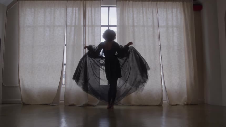 A woman wearing a sheer black dress stands silhouetted against a large window framed by white curtains and then she raises her arms and twirls so the fabric of her skirt fans out around her in a dark circular shape