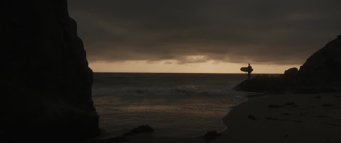 A surfer stands on a beach at sunset