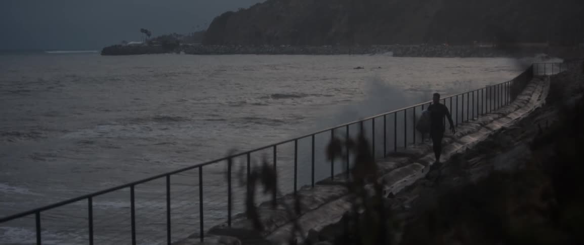 Heavy waves hit the shore line as a man carries a surfboard
