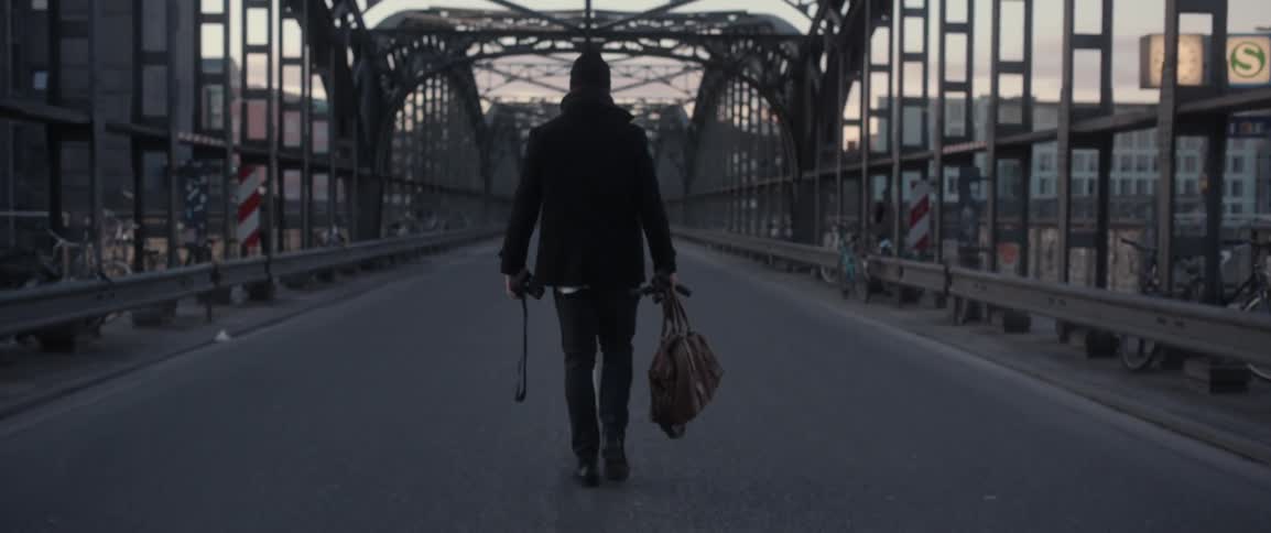 A man carrying a bag and camera walks down the center line of a bridge