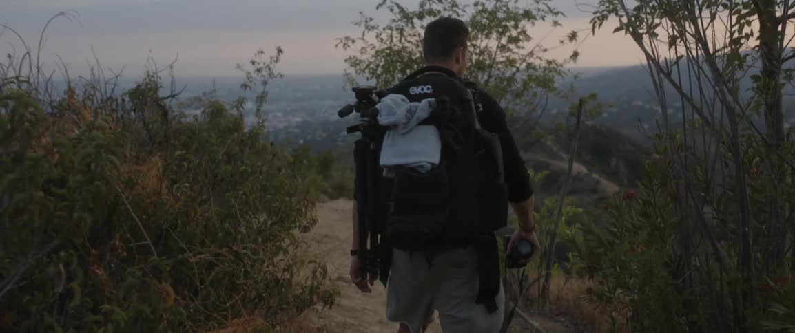 A man wearing a backpack and holding a camera hikes down a path