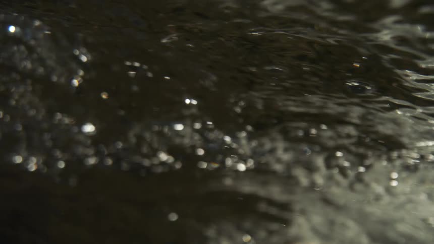 An extreme closeup handheld shot captures the rapid movement of dark turbulent water emphasizing the texture of white bubbles and splashing droplets