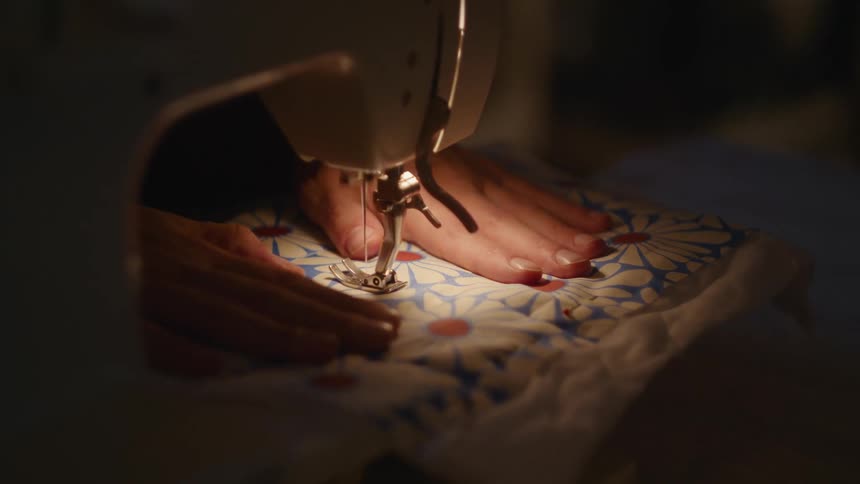 A tight closeup shows the needle and presser foot of a sewing machine stitching across a floralpatterned piece of fabric brightly illuminated by a small lamp as a persons hands carefully guide the material during the quilting process