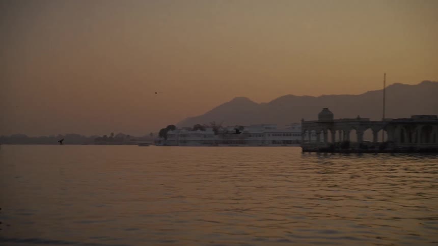 The video is a wide shot across a body of water at dusk featuring a soft orange and brown color palettethe camera pans slowly across the water revealing a complex of historic white buildings potentially a palace or hotel and distant mountains silh