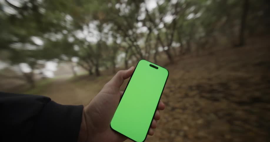 The video shows a closeup of a hand holding a modern smartphone with a bright green screen which is typically used for chroma key editing the camera is moving quickly blurring the natural background which appears to be a forest or wooded area