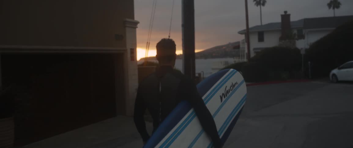 A man carries a surfboard on a road at dawn