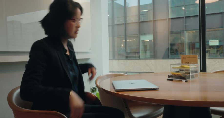 A woman dressed in a black suit walks into a modern bright office room sits down at a round wooden table places her silver laptop on the surface and opens it to begin working