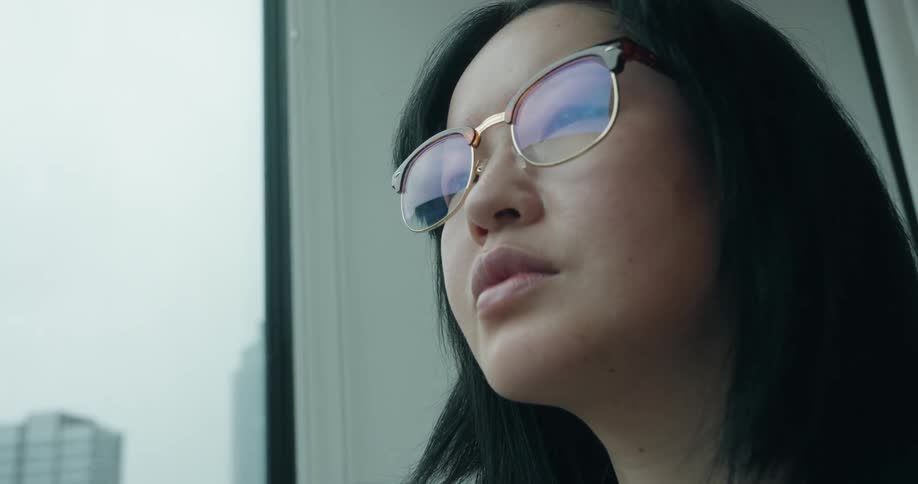 A pensive woman wearing glasses gazes out a high rise window at the city skyline