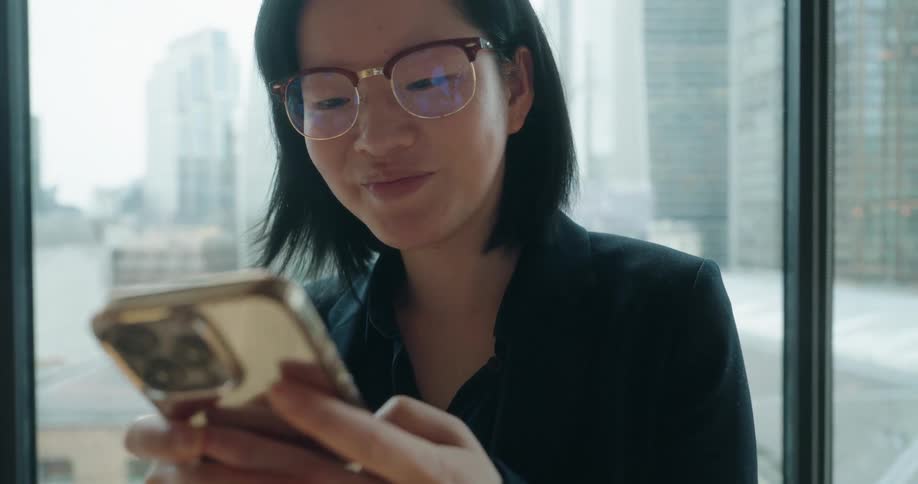 A woman in a black suit and glasses is looking at her phone while standing in front of a window overlooking a cityscapeshe seems pleased or amused by what she is reading on the device smiling slightly as she scrolls