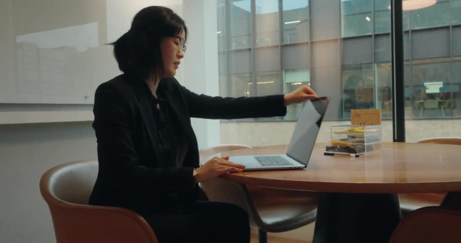 An asian woman wearing a black suit and glasses walks into a room sits behind a chair next to a coffee table places her laptop on the table and begins typing
