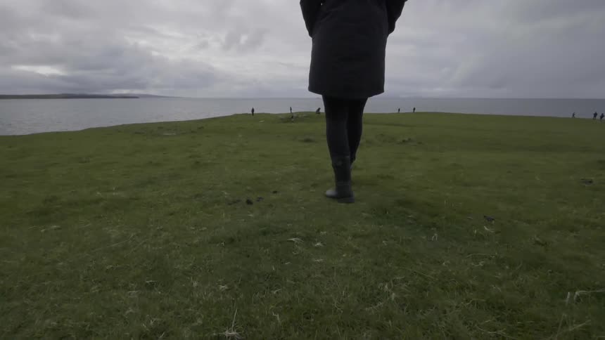 A woman walks on a grassy field overlooking a lake