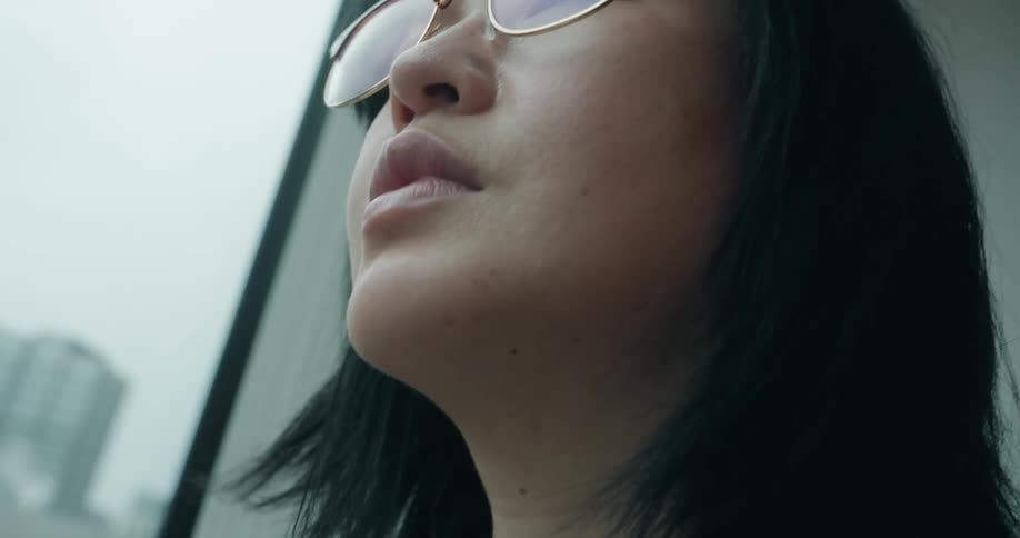 A woman adjusts her glasses as she stands next to a window looking out at the city