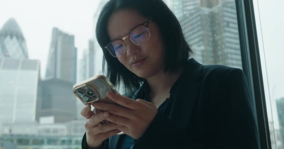 Woman standing near a window with a view of downtown buildings as she texts on her phone