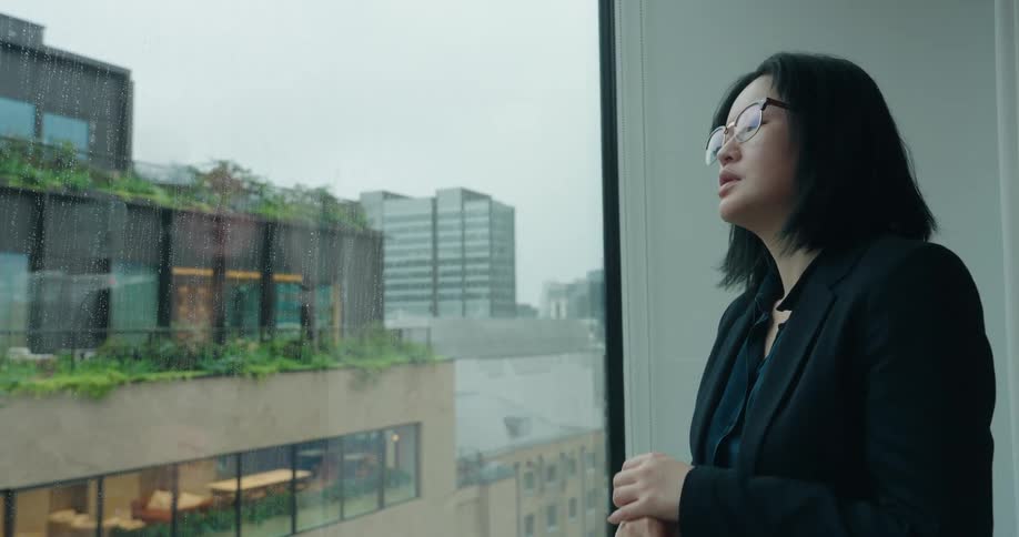 A static medium shot captures a woman in a blazer checking her watch near a large window before looking out at the rainy city skyline and adjusting her glasses