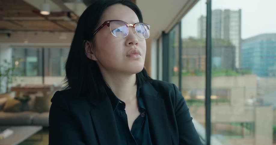 A serious asian businesswoman in a black blazer stands by a large window overlooking a city putting on her glasses and looking into the distance