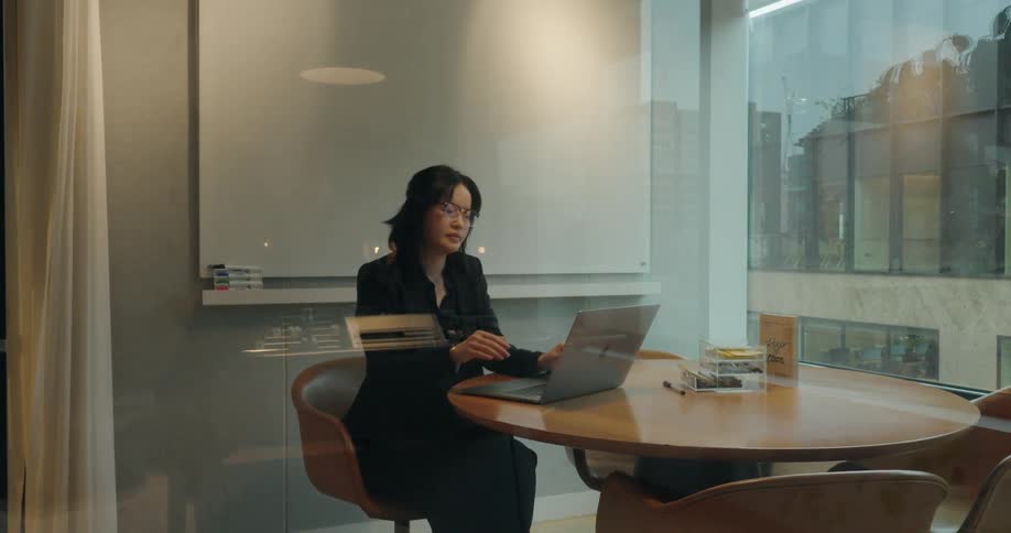 A woman in a black suit enters a glass walled office sits at a round wooden table and opens her laptop to begin working