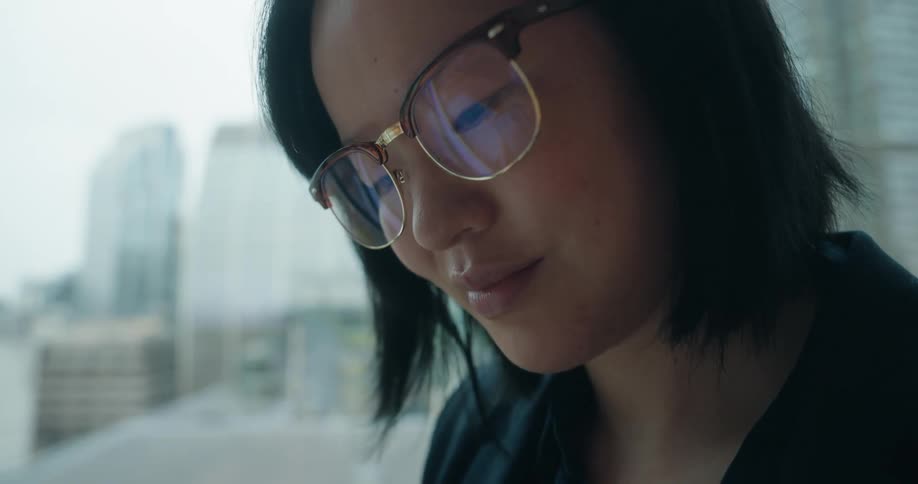 Woman with glasses using a phone in front of windows with buildings in the background