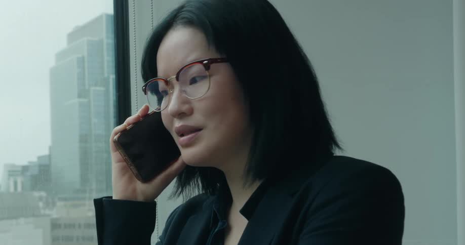Woman stands near a wall next to a window as she talks on her phone