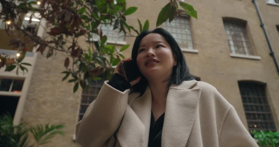 A young asian woman in a pale coat stands outside under trees talking seriously on a mobile phone against a brick building background