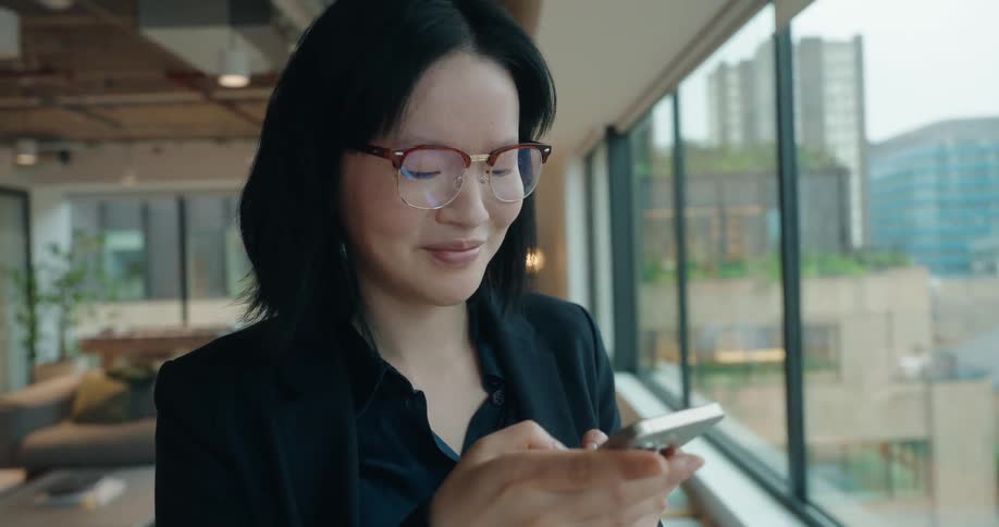 A young asian woman wearing glasses stands beside a glass window inside an office building at midday smiling as she types on her phone