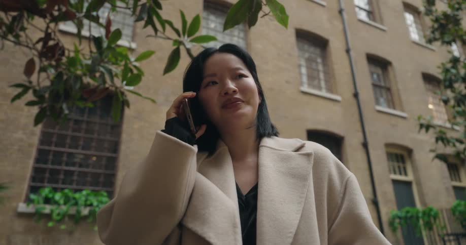 An asian woman wearing a jacket stands in the middle of the street at midday smiling as she makes a phone call