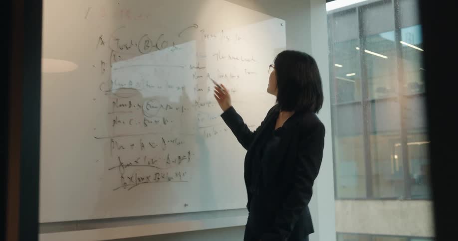 This video shows a professional woman with short dark hair and glasses dressed in a black suit giving a presentation in a modern officeshe stands in front of a whiteboard filled with handwritten notes and diagrams pointing and gesturing towards the