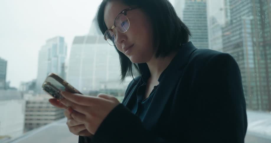 Woman in executive attire looks at smartphone near windows overlooking buildings in london