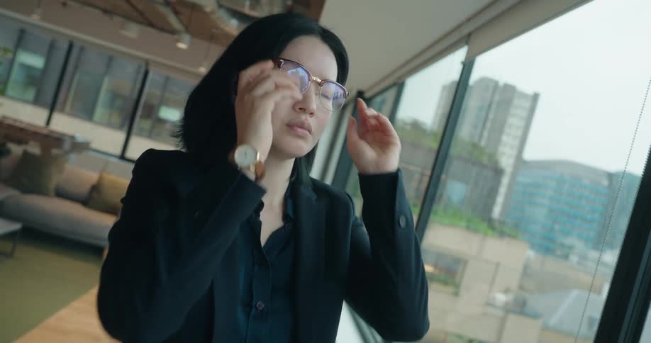 A professionally dressed woman stands by a large floortoceiling window in a modern office lounge area looking out at the city view then raises her eyeglasses and puts them on settling into a focused gaze