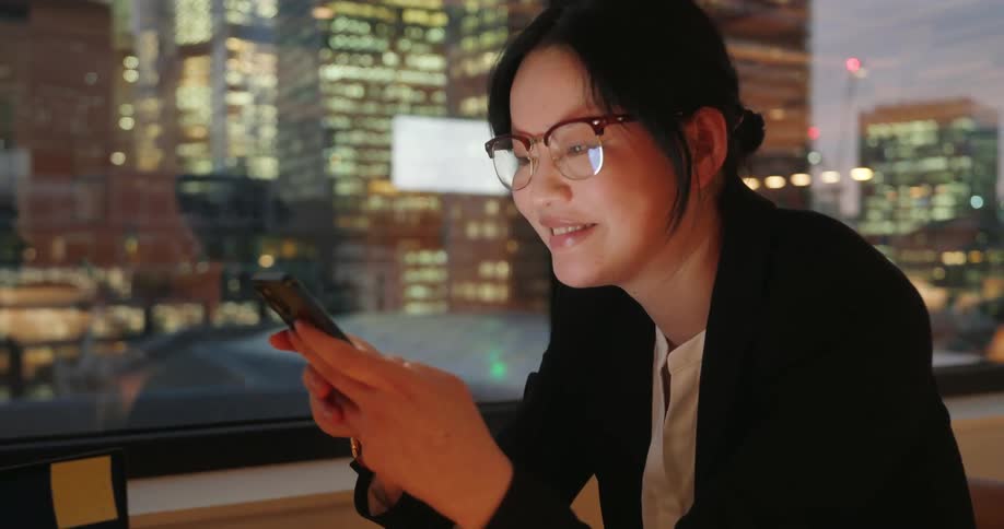 Woman using her phone near the window with illuminated buildings in the background