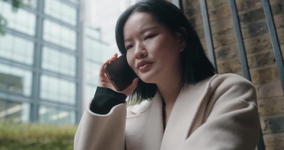 A young woman wearing a jacket stands beside a building at midday making a phone call