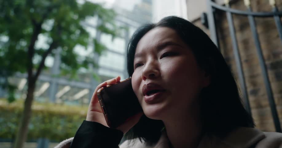 Woman with black hair and beige coat talks on phone near modern buildings and urban greenery