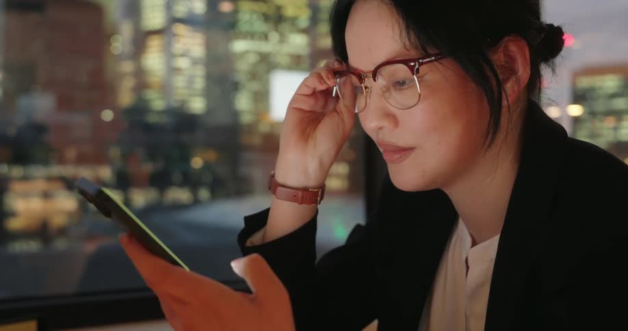 Woman adjusts her glasses while looking at her phone in front of illuminated buildings