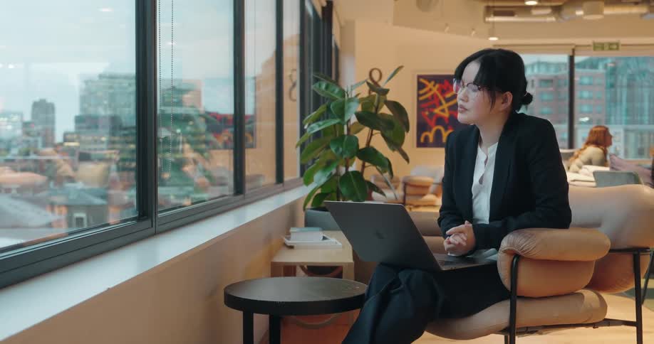 A young asian woman sits beside a window in a public office area typing on a laptop resting on her lap