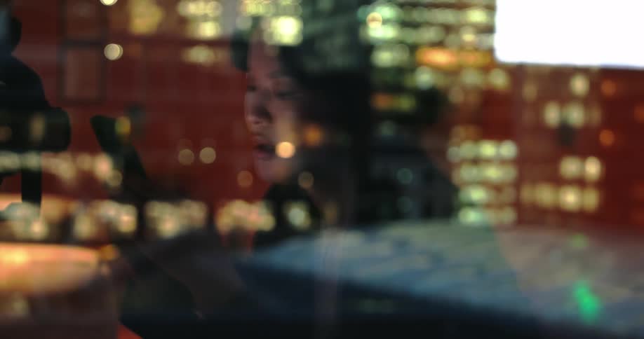 Woman using her phone reflected in the window with city lights in the background