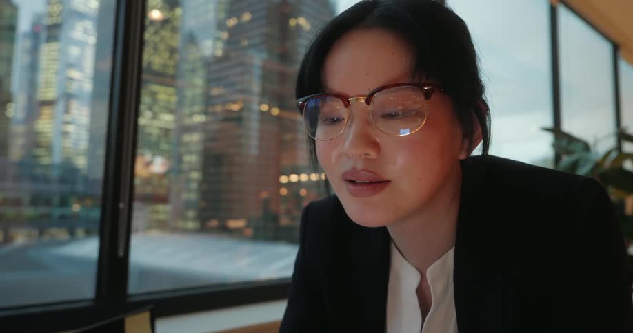 A focused woman in a black blazer and glasses works at a table by a large window with a view of illuminated city buildings at night
