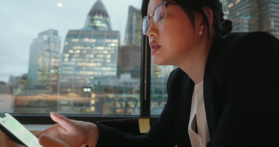 Young asian woman wearing glasses reading on a smartphone on her hand next to a window showing a metropolitan area outdoors during day time