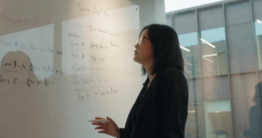 A young asian woman in a black suit and glasses writes on a whiteboard inside a conference room
