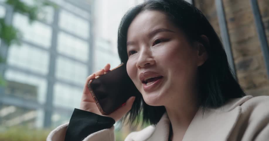 Woman with black hair and light coat talks on phone near urban building with large windows