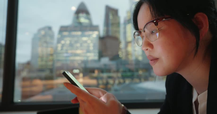 Young asian woman texting on her phone with a nervous facial expression sitting next to a window showing a metropolitan area indoors during day time