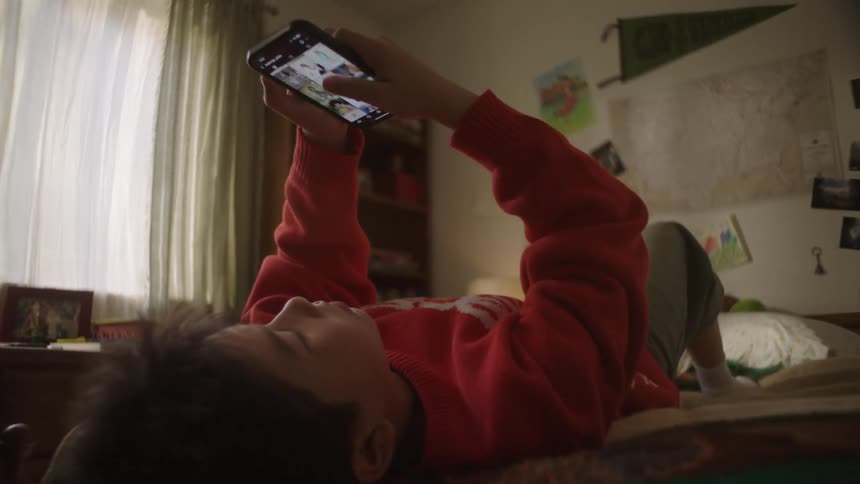 A child lies on his back on a bed in a sunlit room holding a smartphone above the head and scrolling through content framed by the dark doorway of the room
