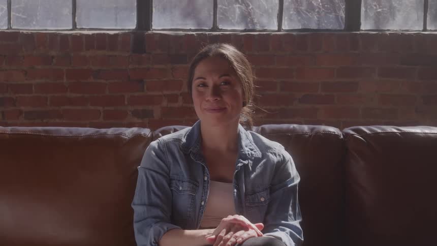 Smiling woman sits on a brown leather couch by a brick wall and large window