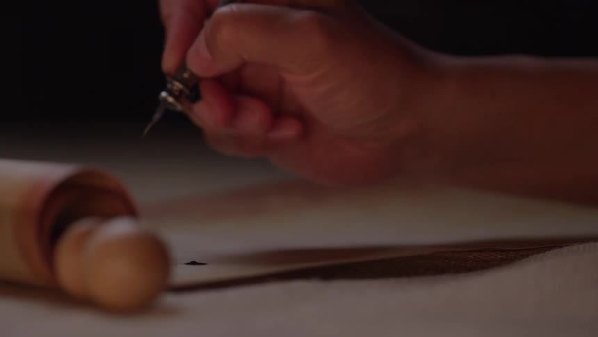 A closeup shot shows a persons hand holding a dip pen and carefully writing on an unrolled scroll with a wooden handle resting outoffocus in the foreground