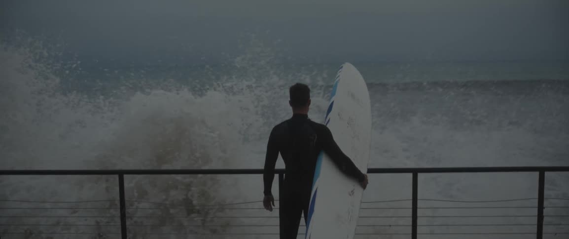 Man holding surfboard standing on walkway watching crashing waves