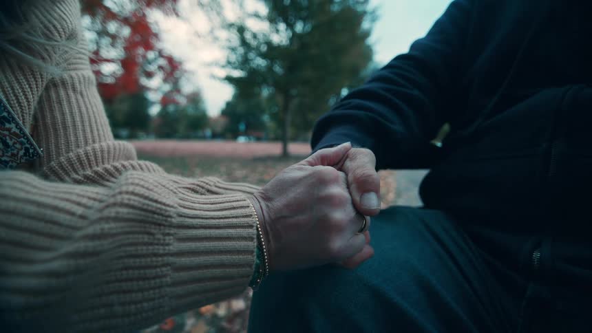 The clasped hands of a man and a woman who are seated closely together outdoors in a park with trees and scattered autumn colors visible in the softfocus background