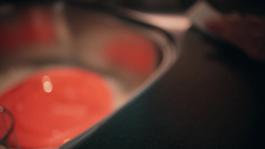 A persons hand using a white paper towel to wipe down a dark surface with the nozzle of a red spray bottle visible in the top right corner and a blurry orange reflection dominating the background