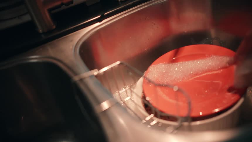 A persons hands rinsing food scraps and residue from a white bowl and an orange plate in a double stainless steel sink with the water and dish soap creating bubbles around the dishes