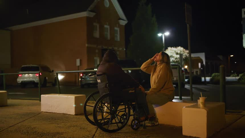 A man in a wheelchair and a woman sitting next to him on a low wall are talking outside a building at night in a dimly lit parking lot